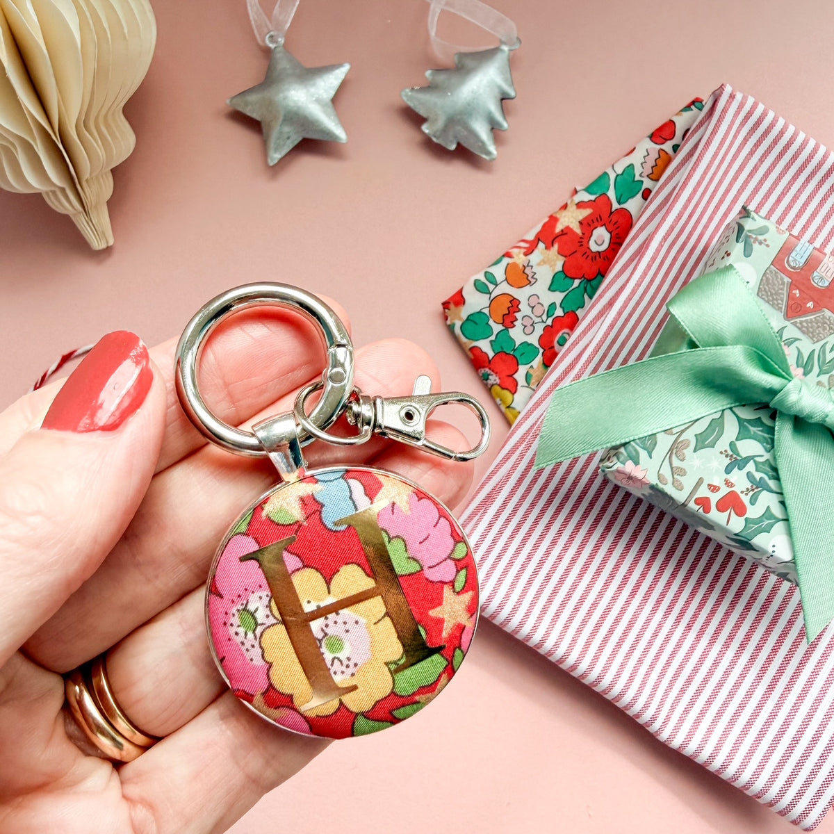 Personalsied gift tag which doubles up as a keyring, made with Liberty of London red floral and star fabric, with an H in gold print, on a Christmas flatlay