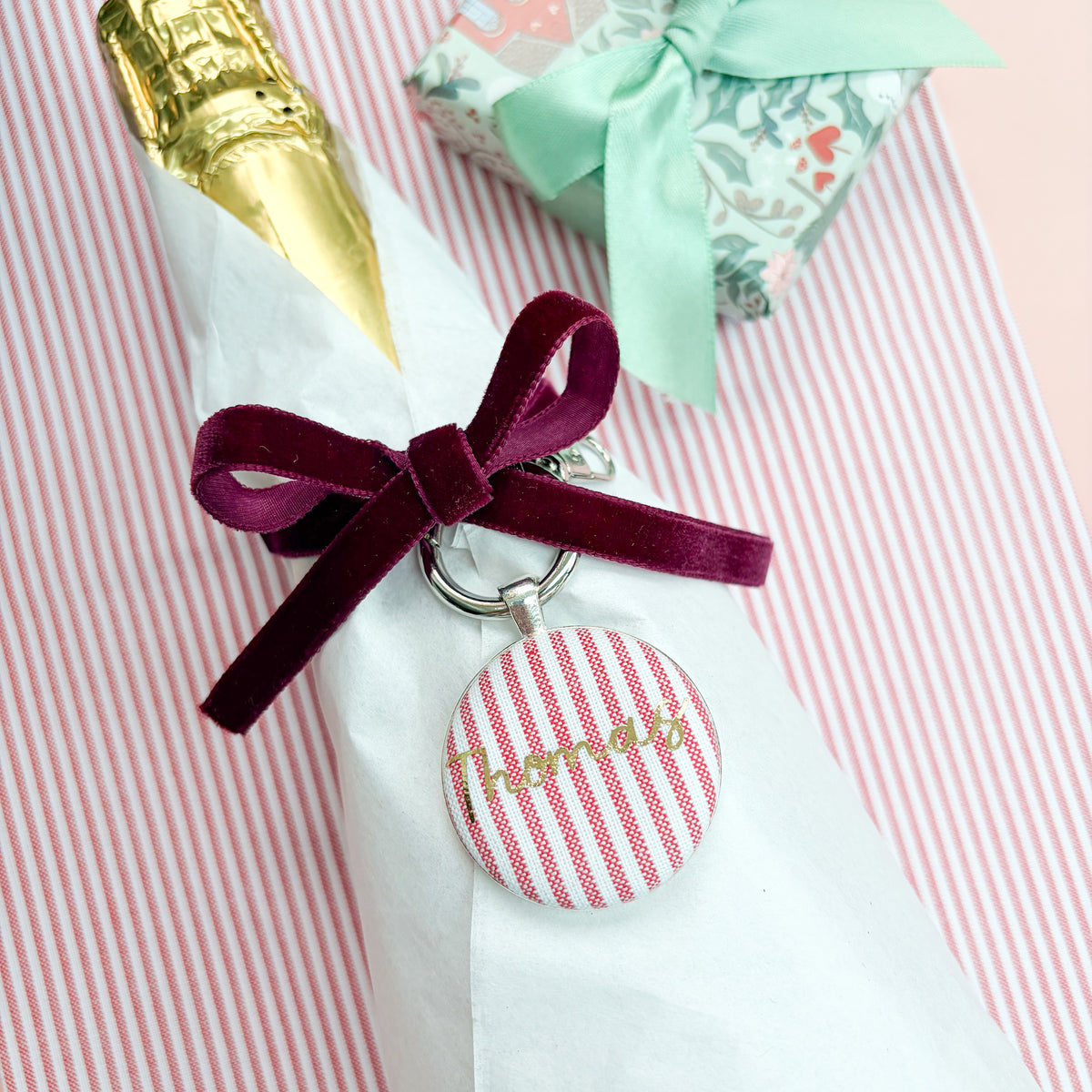 Personalised Christmas Gift Tag and Keyring, made with festive red striped fabric, printed with Thomas in gold foil, set in a keyring, thread on a burgundy velvet ribbon, tied around a bottle of wine and set on a Christmas red striped fabric