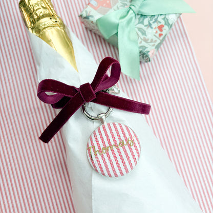 Personalised Christmas Gift Tag and Keyring, made with festive red striped fabric, printed with Thomas in gold foil, set in a keyring, thread on a burgundy velvet ribbon, tied around a bottle of wine and set on a Christmas red striped fabric
