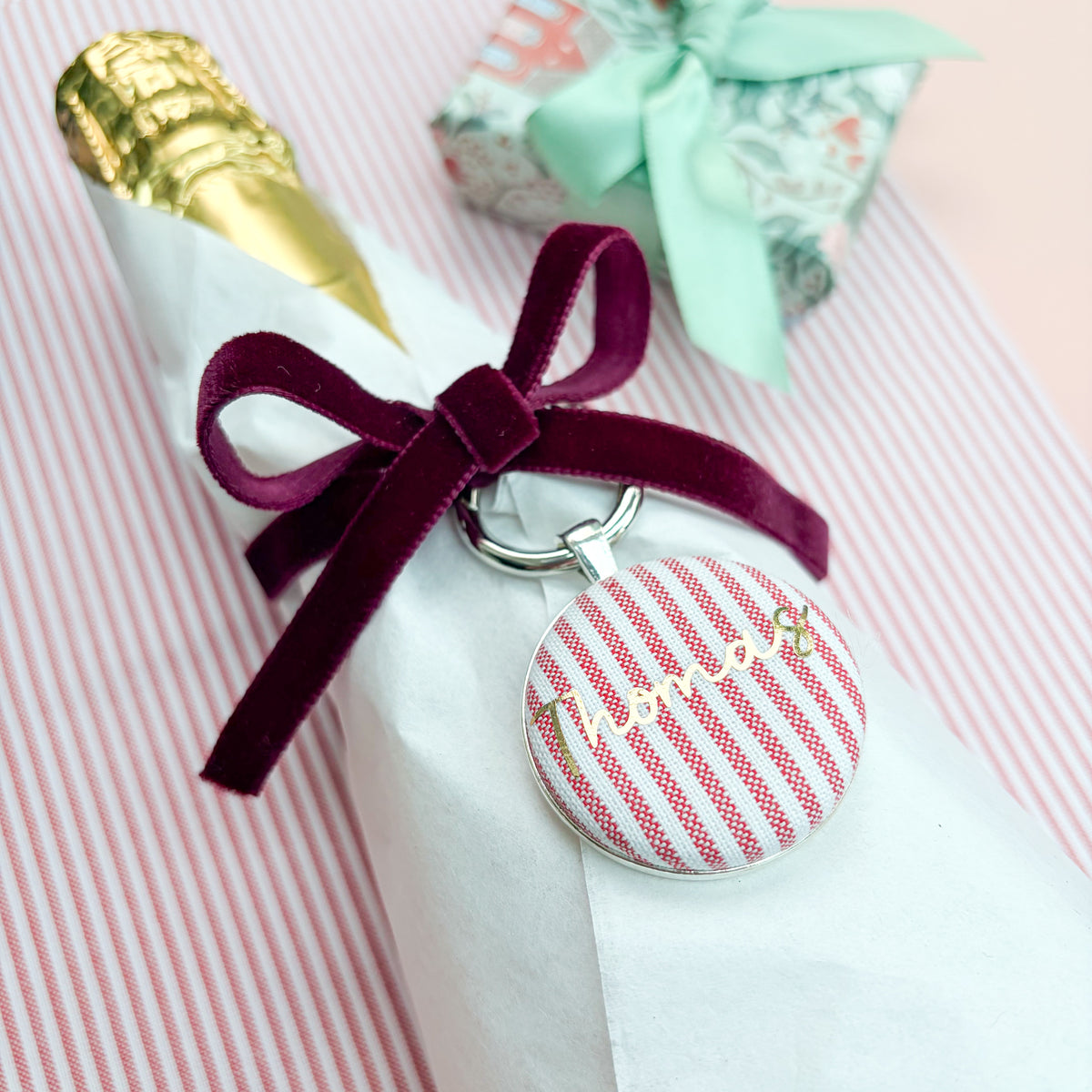 Personalised Christmas Gift Tag and Keyring, made with festive red striped fabric, printed with Thomas in gold foil, set in a keyring, thread on a burgundy velvet ribbon, tied around a bottle of wine and set on a Christmas red striped fabric