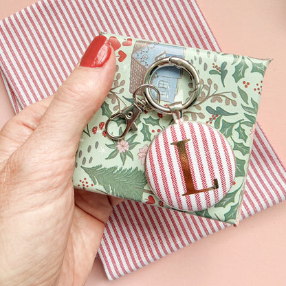 Personalised Christmas Gift Tag and Keyring, made with festive red striped fabric, printed with the initial L in gold foil, set in a keyring, with a small gift and set on a Christmas red striped fabric