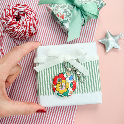 Personalised Christmas Gift Tag and Keyring, made with white Liberty floral and star fabric, printed with AW in gold foil, set in a keyring, thread on a white velvet ribbon, tied around a small gift and set on a Christmas flatlay