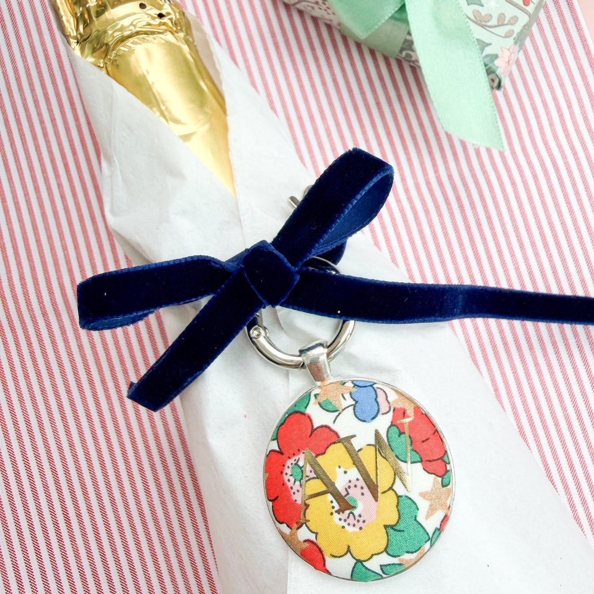 Personalised Christmas Gift Tag and Keyring, made with white Liberty floral and star fabric, printed with AW in gold foil, set in a keyring, thread on a blue velvet ribbon, tied around a bottle of wine and set on a Christmas red striped fabric