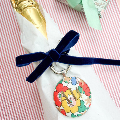 Personalised Christmas Gift Tag and Keyring, made with white Liberty floral and star fabric, printed with AW in gold foil, set in a keyring, thread on a blue velvet ribbon, tied around a bottle of wine and set on a Christmas red striped fabric