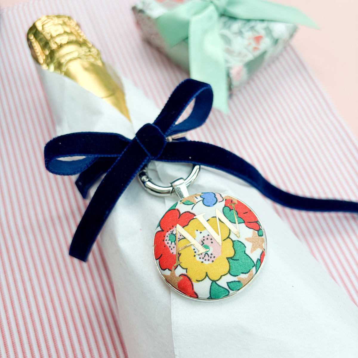 Personalised Christmas Gift Tag and Keyring, made with white Liberty floral and star fabric, printed with AW in gold foil, set in a keyring, thread on a blue velvet ribbon, tied around a bottle of wine and set on a Christmas red striped fabric
