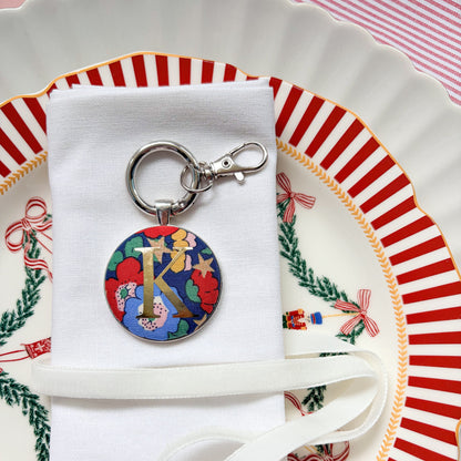 Initial Christmas table place setting, made with blue floral Liberty fabric, printed with a gold K, set in a keyring, displayed on a white napkin and Christmas plates