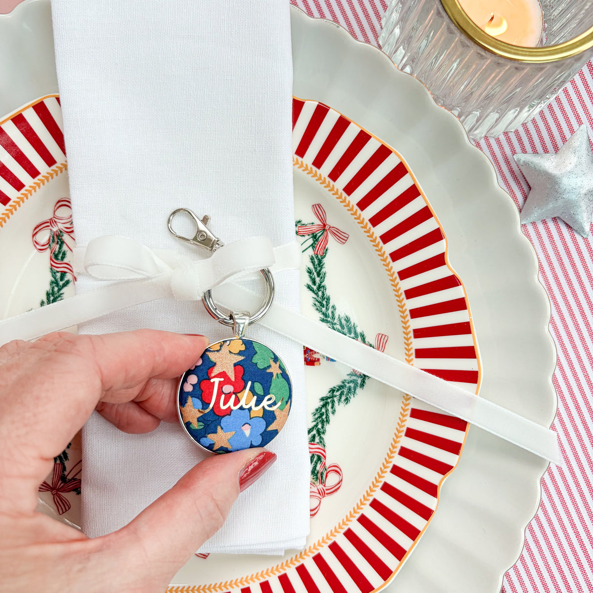 Personalised Christmas Place Setting Keyring, made with Liberty blue floral fabric, printed with Julie in gold, set in a keyring, thread on a velvet ribbon, tied around a napkin, on Christmas plates
