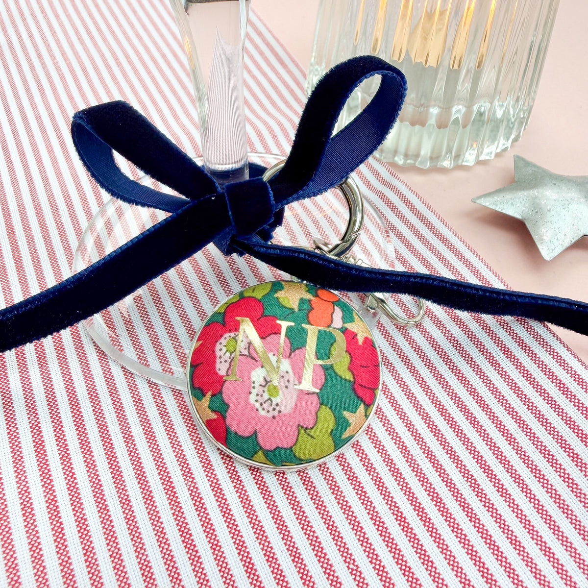Personalised initial table place setting on a keyring, with green floral fabric and gold initials, thread on a blue velvet ribbon and tied around a wine glass, set on red striped fabric