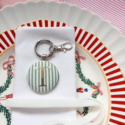 Personalised name place setting for the Christmas table, made with green striped fabric, printed with a T in gold, set in a silver plated keyring, displayed on a white napkin on a Christmas plate