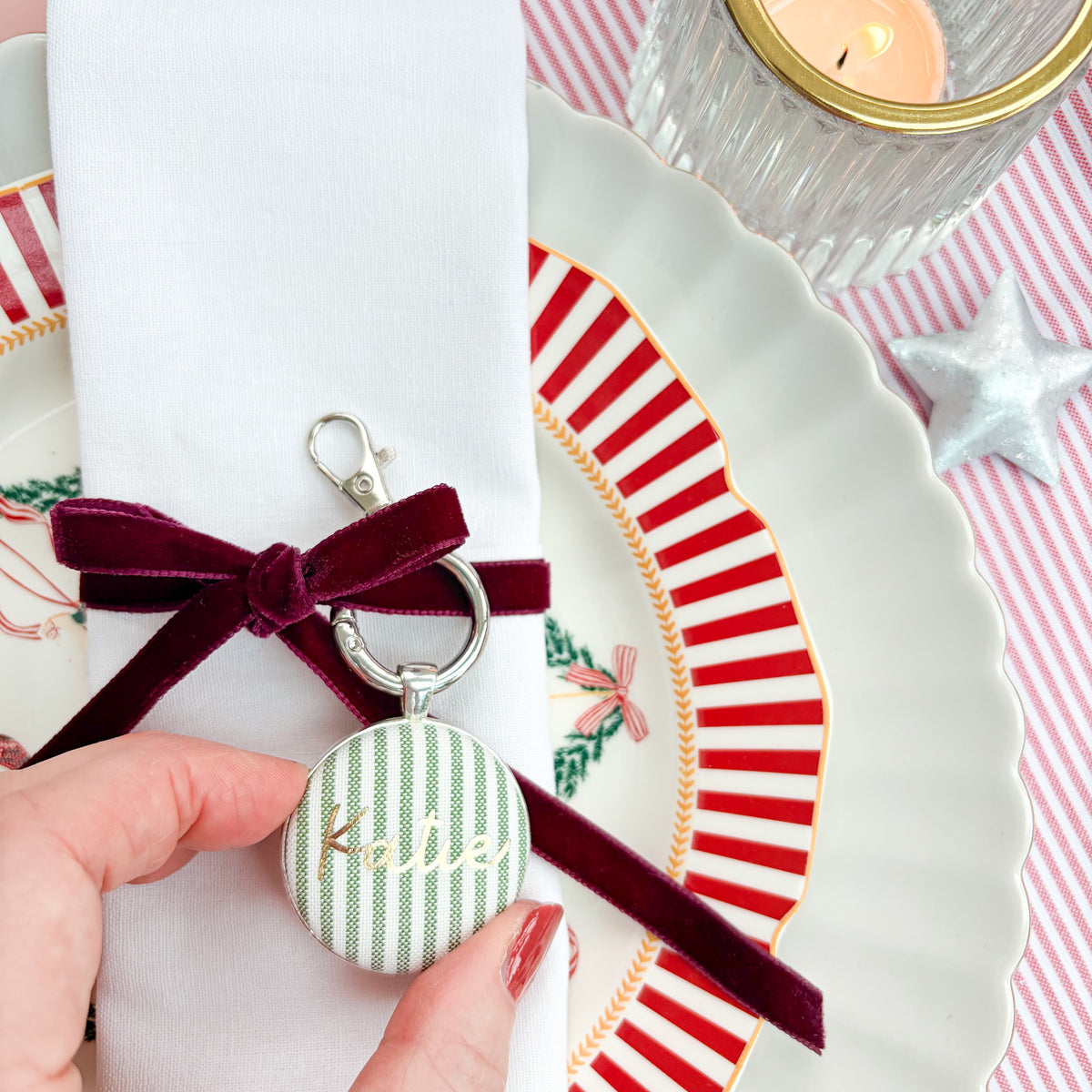Personalised name place setting for the Christmas table, made with green striped fabric, printed with Katie in gold, set in a silver plated keyring, thread on a burgundy velvet ribbon, tied around a white napkin on a Christmas plate