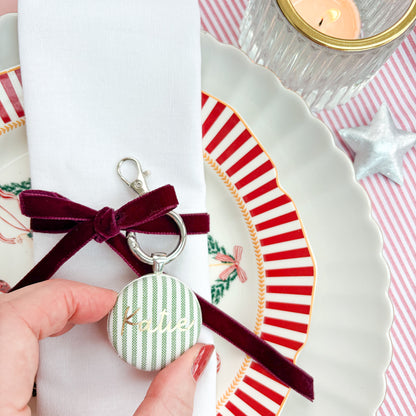 Personalised name place setting for the Christmas table, made with green striped fabric, printed with Katie in gold, set in a silver plated keyring, thread on a burgundy velvet ribbon, tied around a white napkin on a Christmas plate