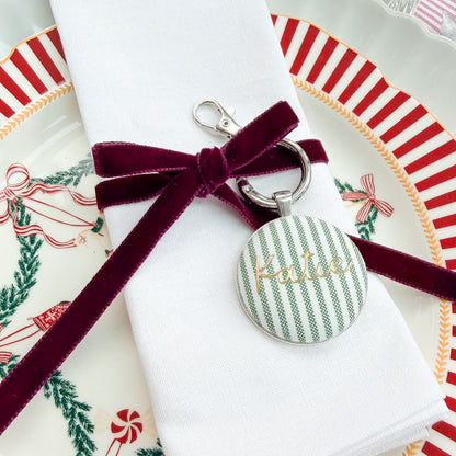 Personalised name place setting for the Christmas table, made with green striped fabric, printed with Katie in gold, set in a silver plated keyring, thread on a burgundy velvet ribbon, tied around a white napkin on a Christmas plate