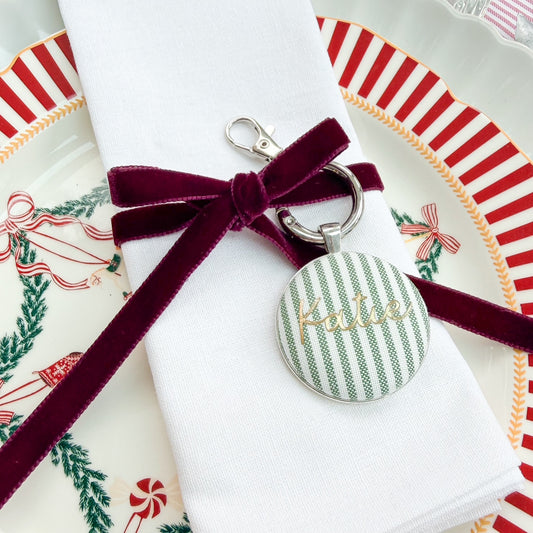 Personalised name place setting for the Christmas table, made with green striped fabric, printed with Katie in gold, set in a silver plated keyring, thread on a burgundy velvet ribbon, tied around a white napkin on a Christmas plate