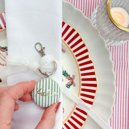 Personalised name place setting for the Christmas table, made with green striped fabric, printed with Katie in gold, set in a silver plated keyring, thread on an ivory white velvet ribbon, tied around a white napkin on a Christmas plate