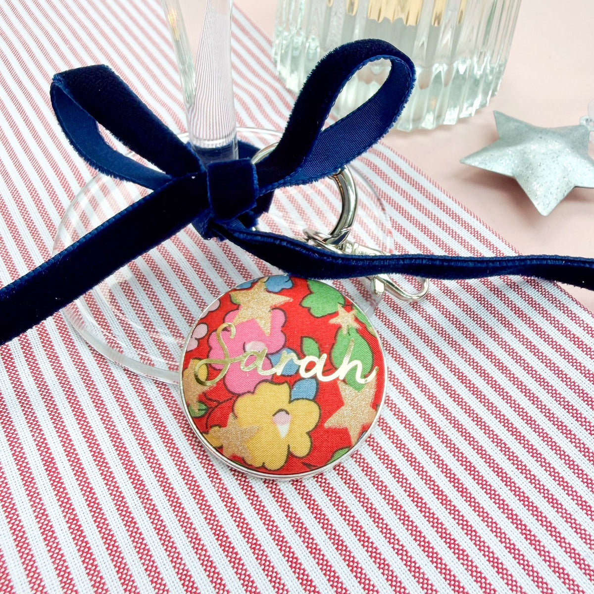 Red floral personalised table place setting, made with Christmas Liberty fabric, the name Sarah in gold print, set in a keyring, thread on a blue velvet ribbon, tied around a wine glass