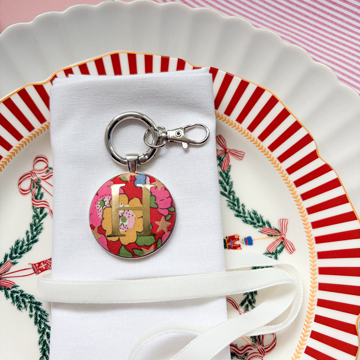 Initial keyring used as a Christmas table place setting, made with red floral Liberty fabric, printed with a gold initial H, set in a keyring, and displayed on a napkin on Christmas plates