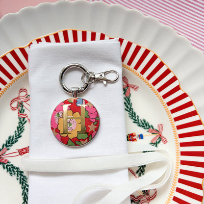 Initial keyring used as a Christmas table place setting, made with red floral Liberty fabric, printed with a gold initial H, set in a keyring, and displayed on a napkin on Christmas plates