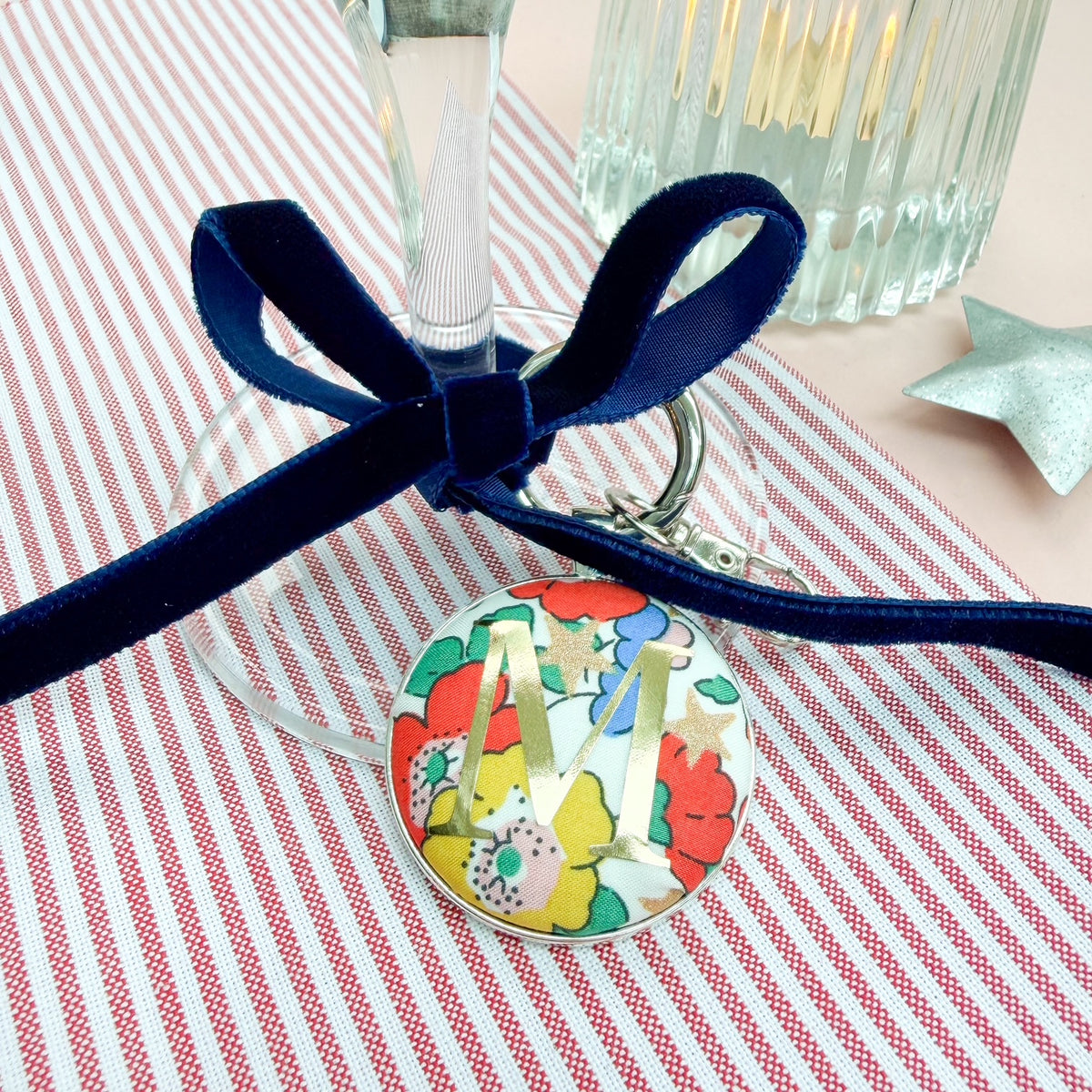 Personalised Christmas place setting keyring, with floral festive fabric and a gold letter M, thread on a blue velvet ribbon, tied in a bow around a wine glass