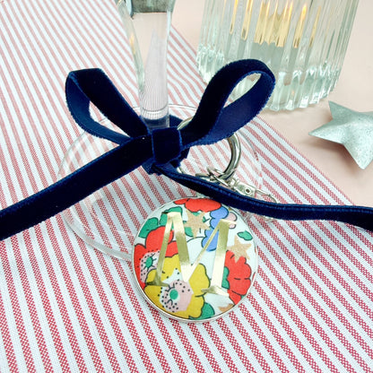 Personalised Christmas place setting keyring, with floral festive fabric and a gold letter M, thread on a blue velvet ribbon, tied in a bow around a wine glass