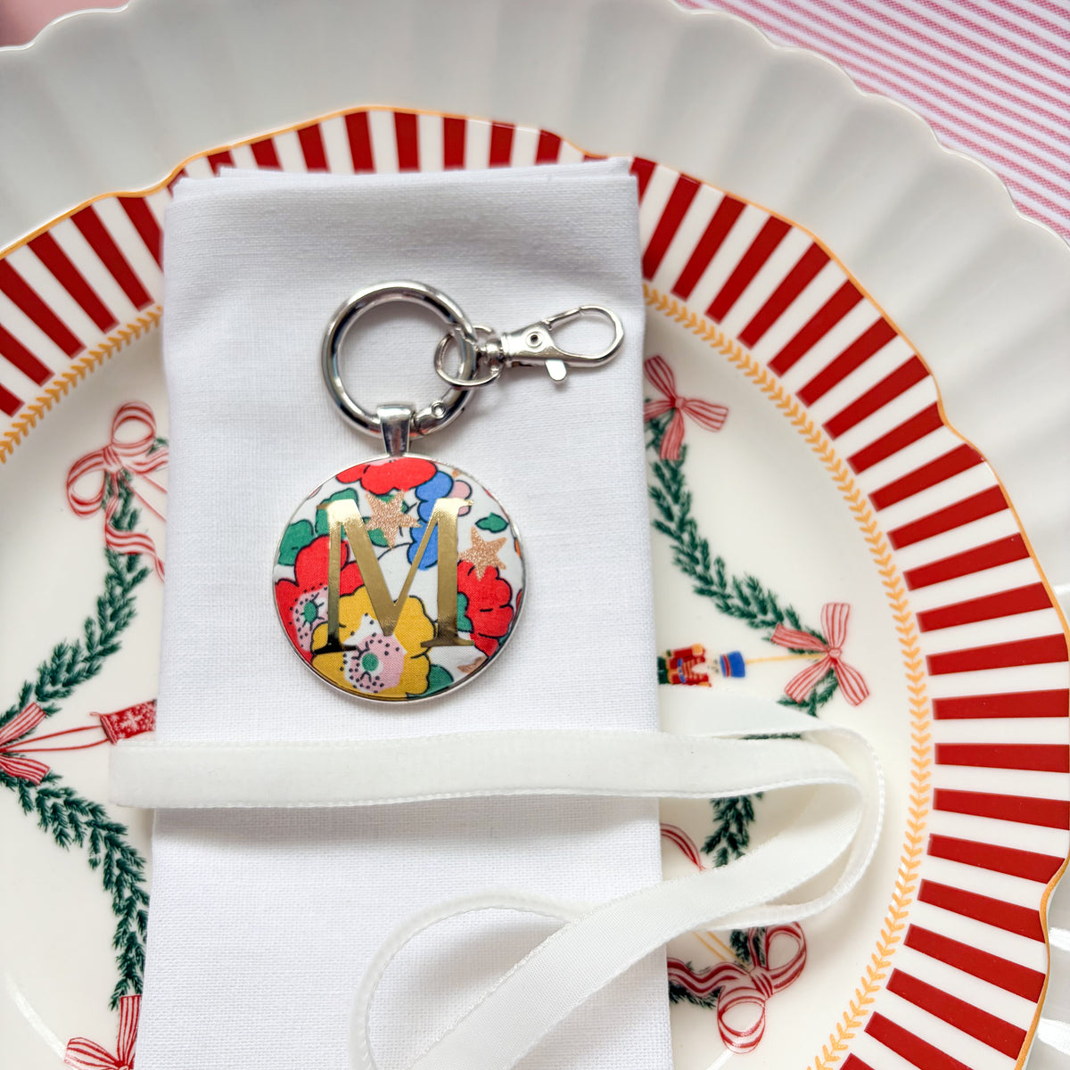 Personalised Christmas table setting made with floral Liberty fabric, printed with a gold M, on a keyring, displayed on a white napkin on Christmas plates