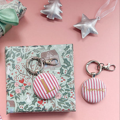 Personalised Christmas table place settings with initials and names, printed in gold on red striped fabric, set in keyrings and displayed on a Christmas flatlay