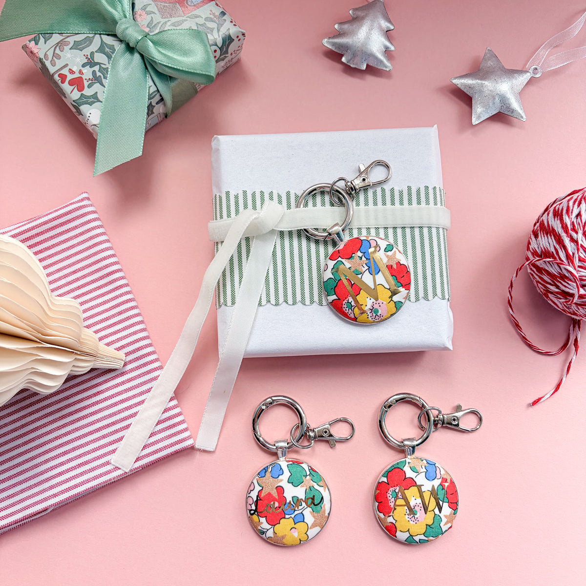 Personalised keyrings which can be used as Christmas table place settings, in multi coloured floral Liberty fabric, with a gold M, tied with velvet ribbon around a gift, and displayed on a  Christmas flatlay