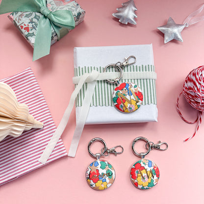 Personalised keyrings which can be used as Christmas table place settings, in multi coloured floral Liberty fabric, with a gold M, tied with velvet ribbon around a gift, and displayed on a  Christmas flatlay