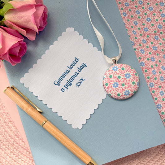 Floral pyjamas memory keepsake, thread on a ribbon, with a fabric message, set on a blue notebook with a pen and rose