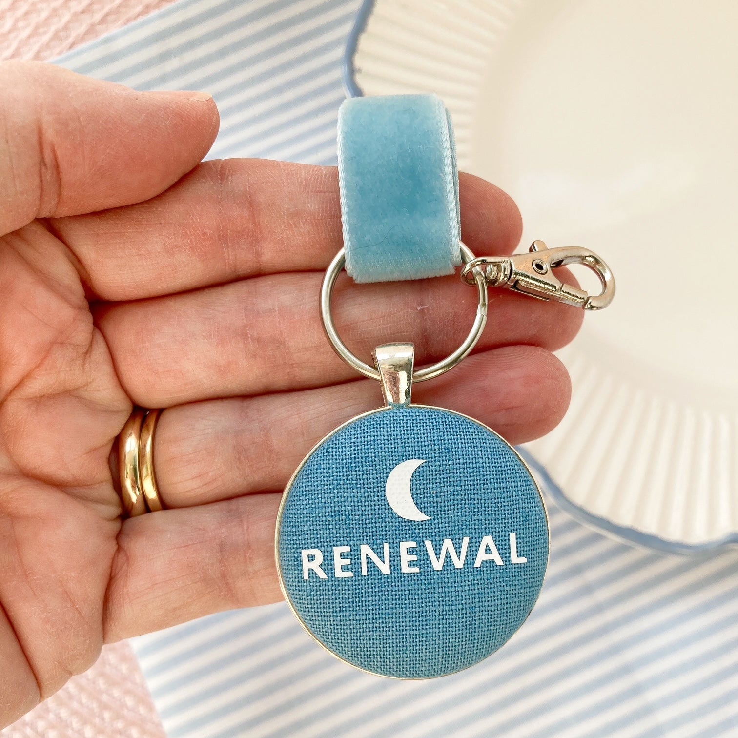 Hand holding a blue key ring and keepsake with 'RENEWAL' text and crescent moon symbol.