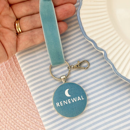 Blue keepsake charm with 'RENEWAL' and a crescent moon, held over a striped fabric background