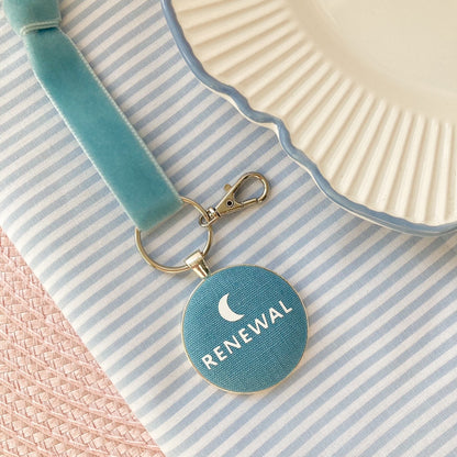 Blue keychain with 'Renewal' text and a new moon printed onto blue fabric, laid on a striped fabric background