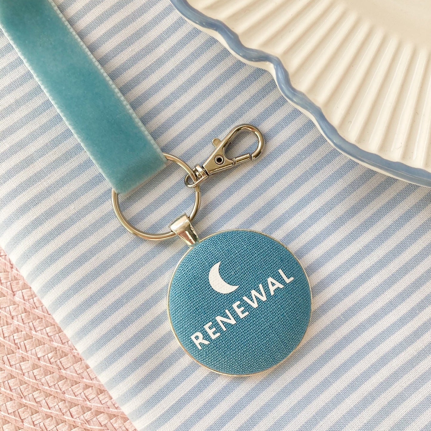 Blue keychain with 'RENEWAL' and moon symbol, on a striped fabric background