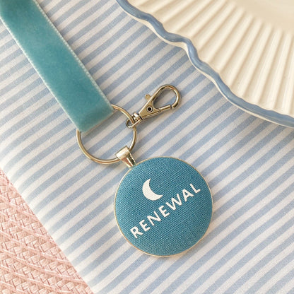 Blue keychain with 'RENEWAL' and moon symbol, on a striped fabric background