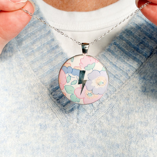 Secret Mantra necklace with a silver lightning printed on a Liberty lilac & pink floral fabric, thread on a silver chain and worn with a pale blue jumper
