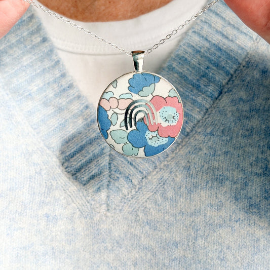 Secret Mantra necklace with a silver rainbow printed on a Liberty blush pink & blue floral fabric, thread on a silver chain and worn with a pale blue jumper