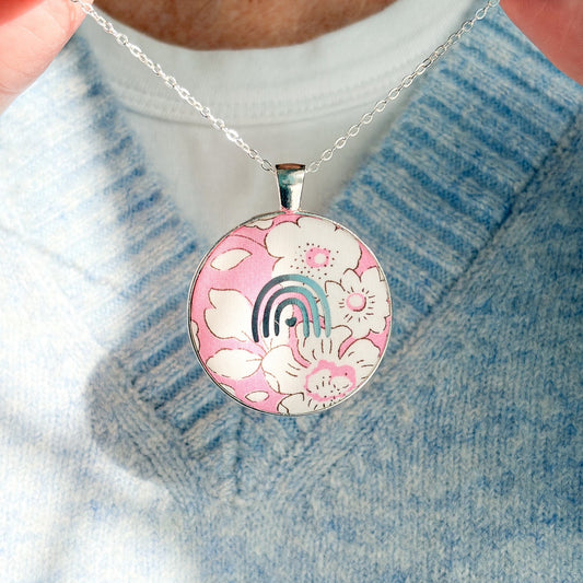 Secret Mantra necklace with a silver rainbow printed on a Liberty pink floral fabric, thread on a silver chain and worn with a pale blue jumper