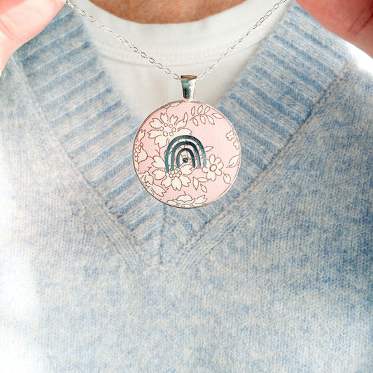 Secret Mantra necklace with a silver rainbow printed on a Liberty pink floral fabric, thread on a silver chain and worn with a pale blue jumper