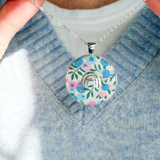 Secret Mantra necklace with a silver rainbow printed on a Liberty blue & pink floral fabric, thread on a silver chain and worn with a pale blue jumper