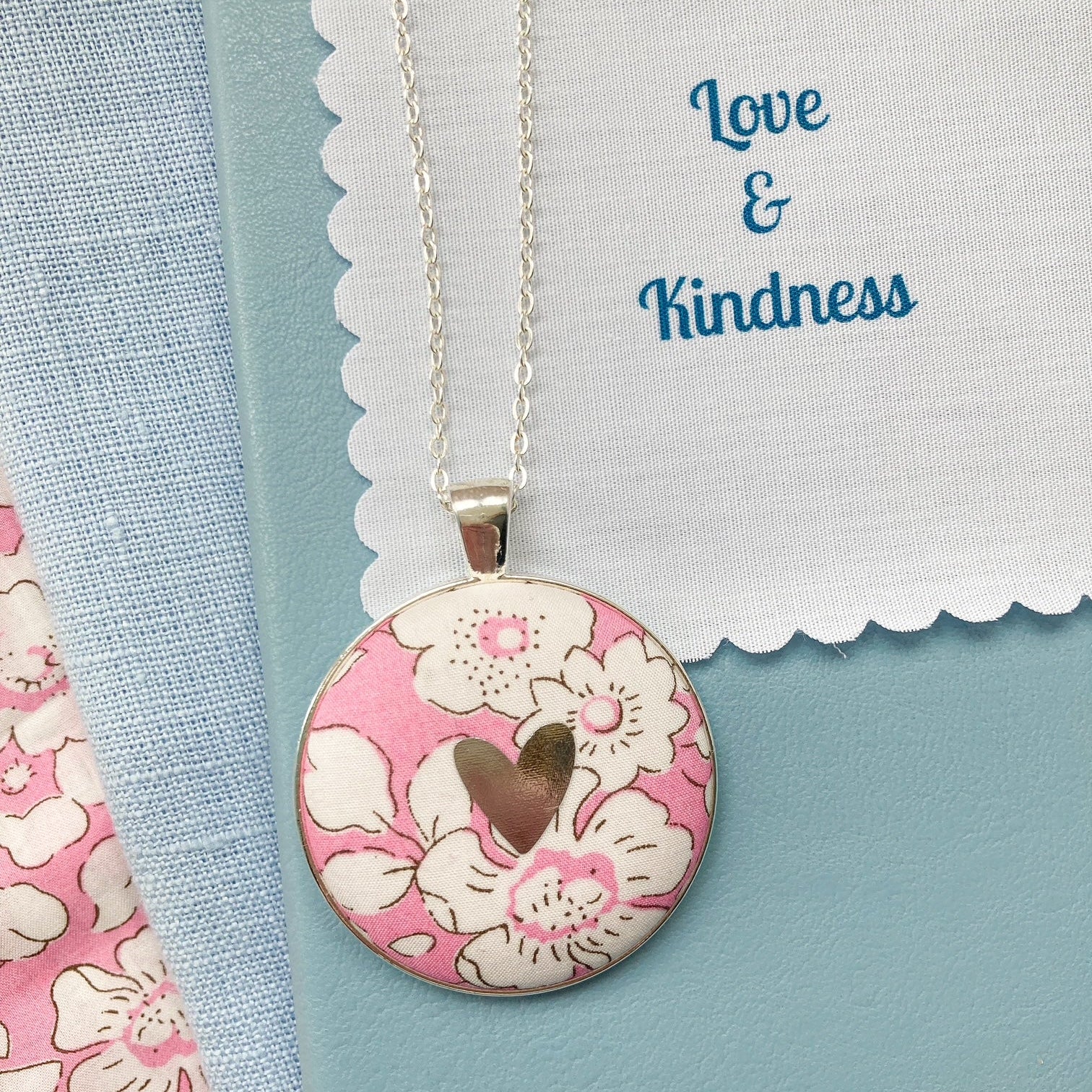 Pink floral fabric necklace with a silver heart on a blue book, with a fabric message saying "Love & Kindness"