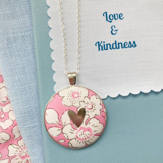 Pink floral fabric necklace with a silver heart on a blue book, with a fabric message saying "Love & Kindness"
