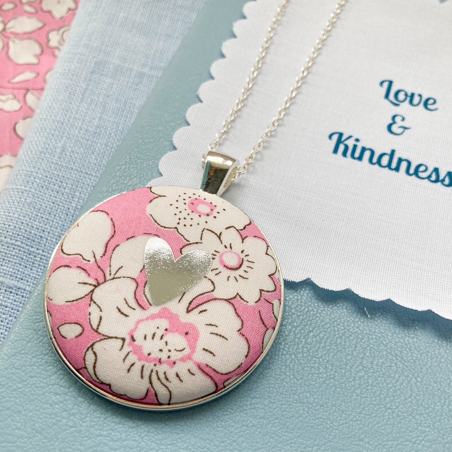 Floral pendant necklace on a light blue background with 'Love & Kindness' fabric message