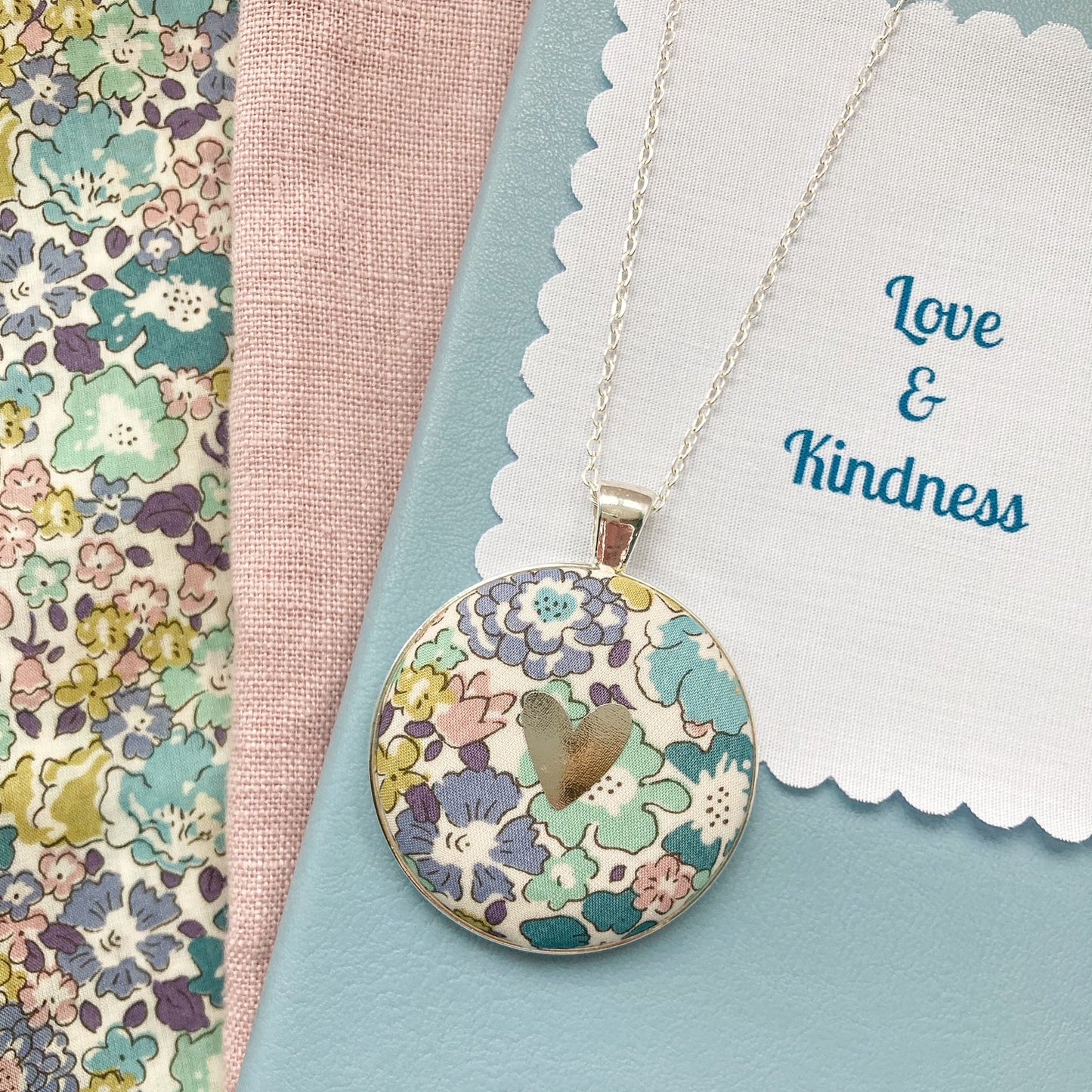 Teal & purple floral necklace with a silver heart and a fabric message saying "Love & Kindness"