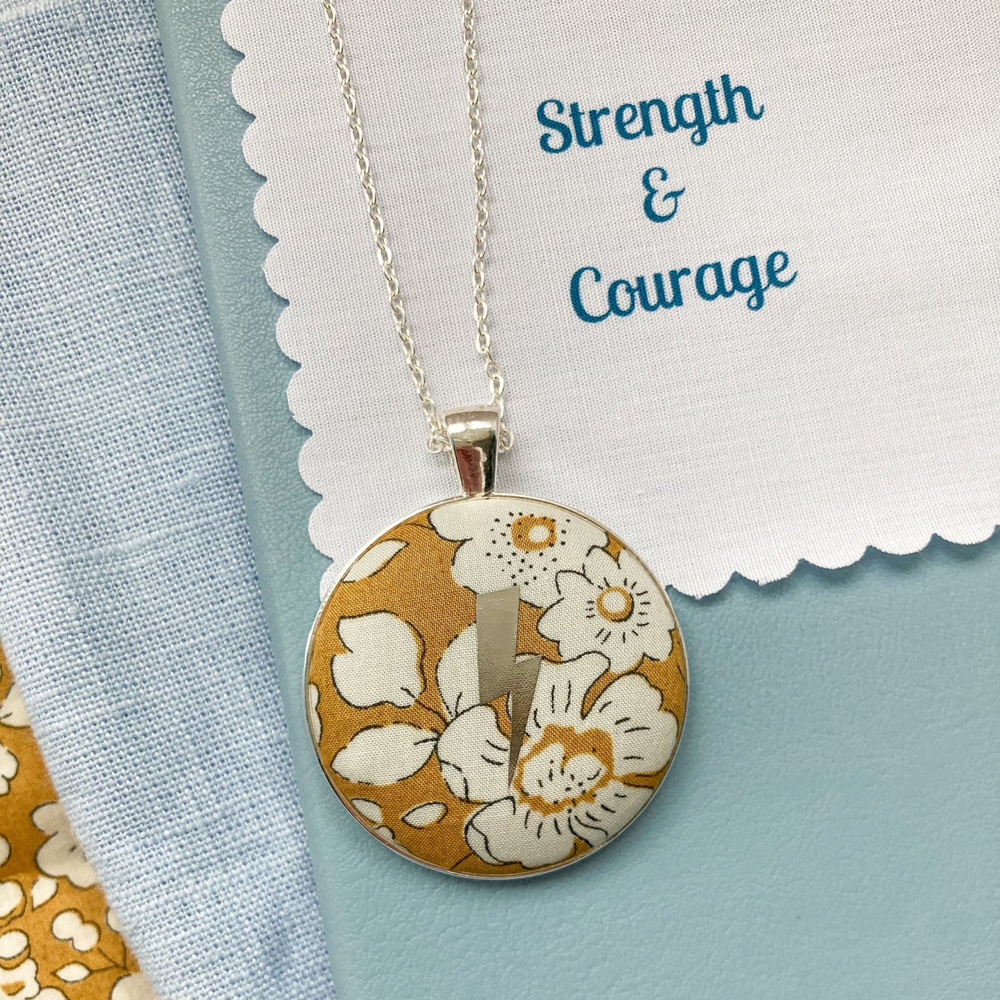 Lightning necklace with floral pendant with fabric message saying 'Strength & Courage', against a light blue background.