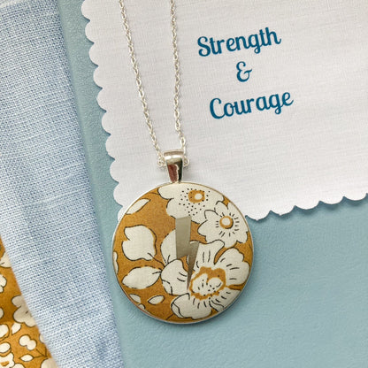 Lightning necklace with floral pendant with fabric message saying 'Strength & Courage', against a light blue background.