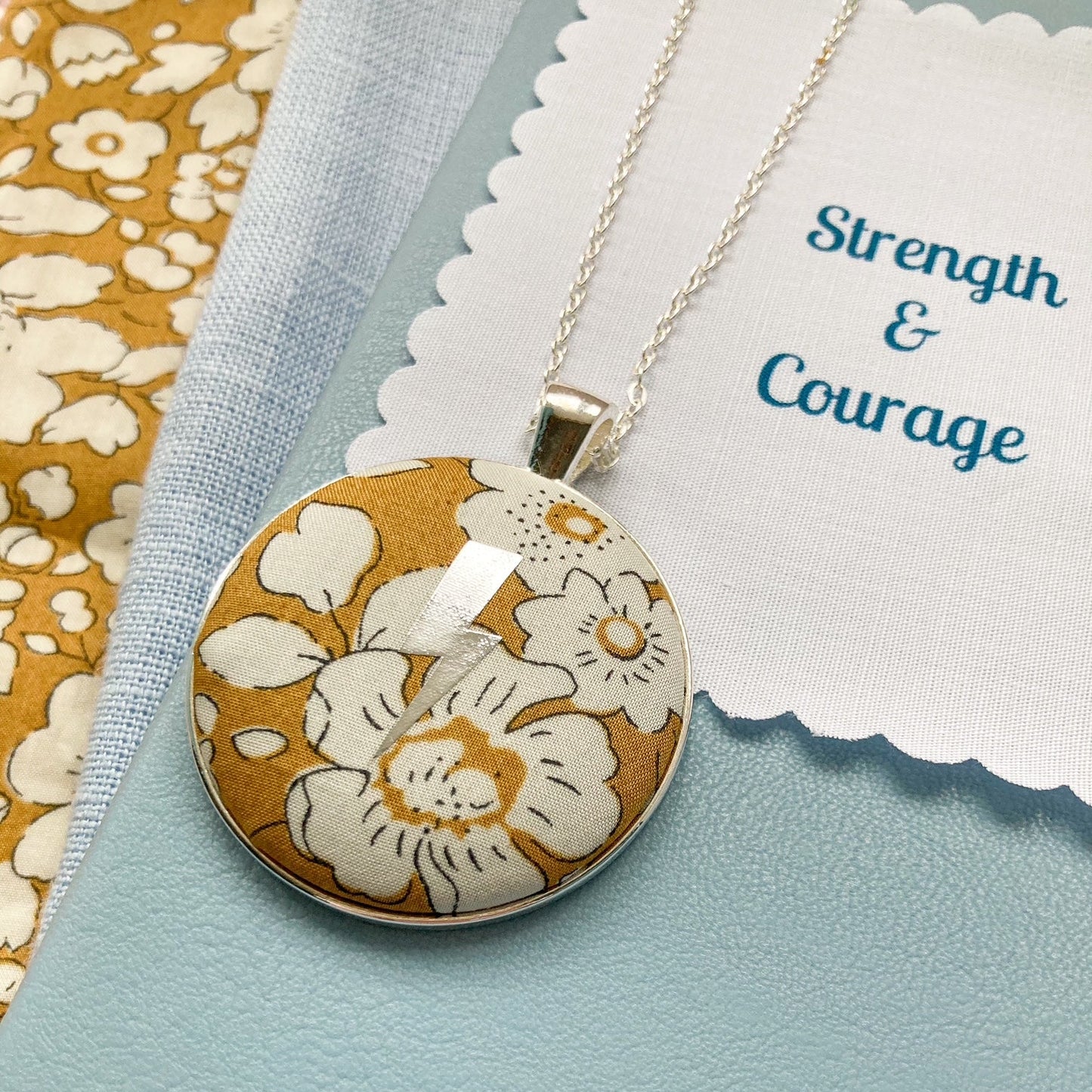 Floral pendant necklace on a blue background with 'Strength & Courage' fabric message