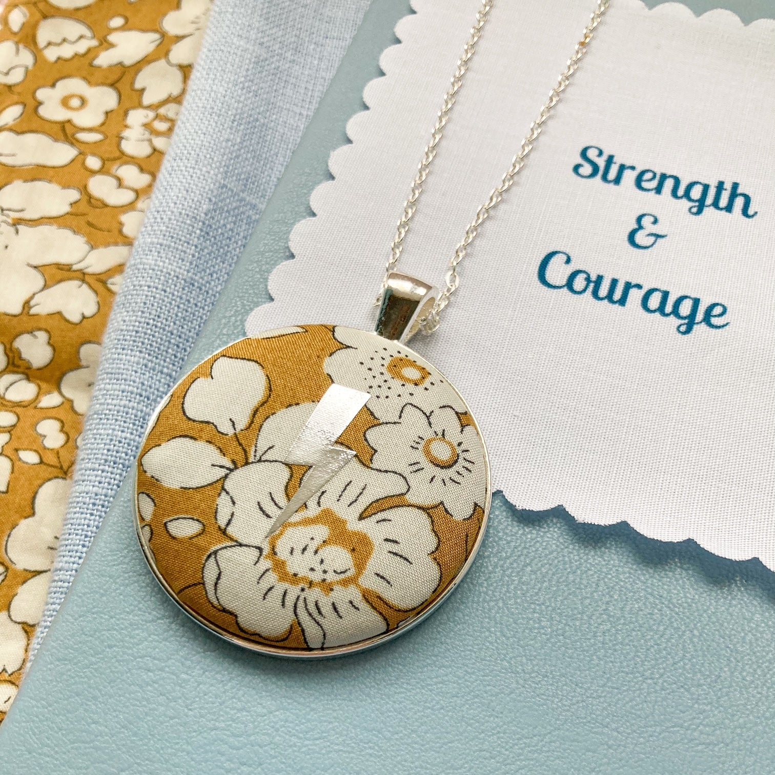 Floral pendant necklace on a blue background with 'Strength & Courage' fabric message