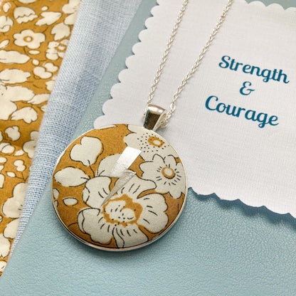 Floral pendant necklace on a blue background with 'Strength & Courage' fabric message