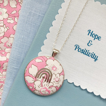 Necklace with a pink floral pendant on a blue fabric background with 'Hope & Positivity' fabric message text.
