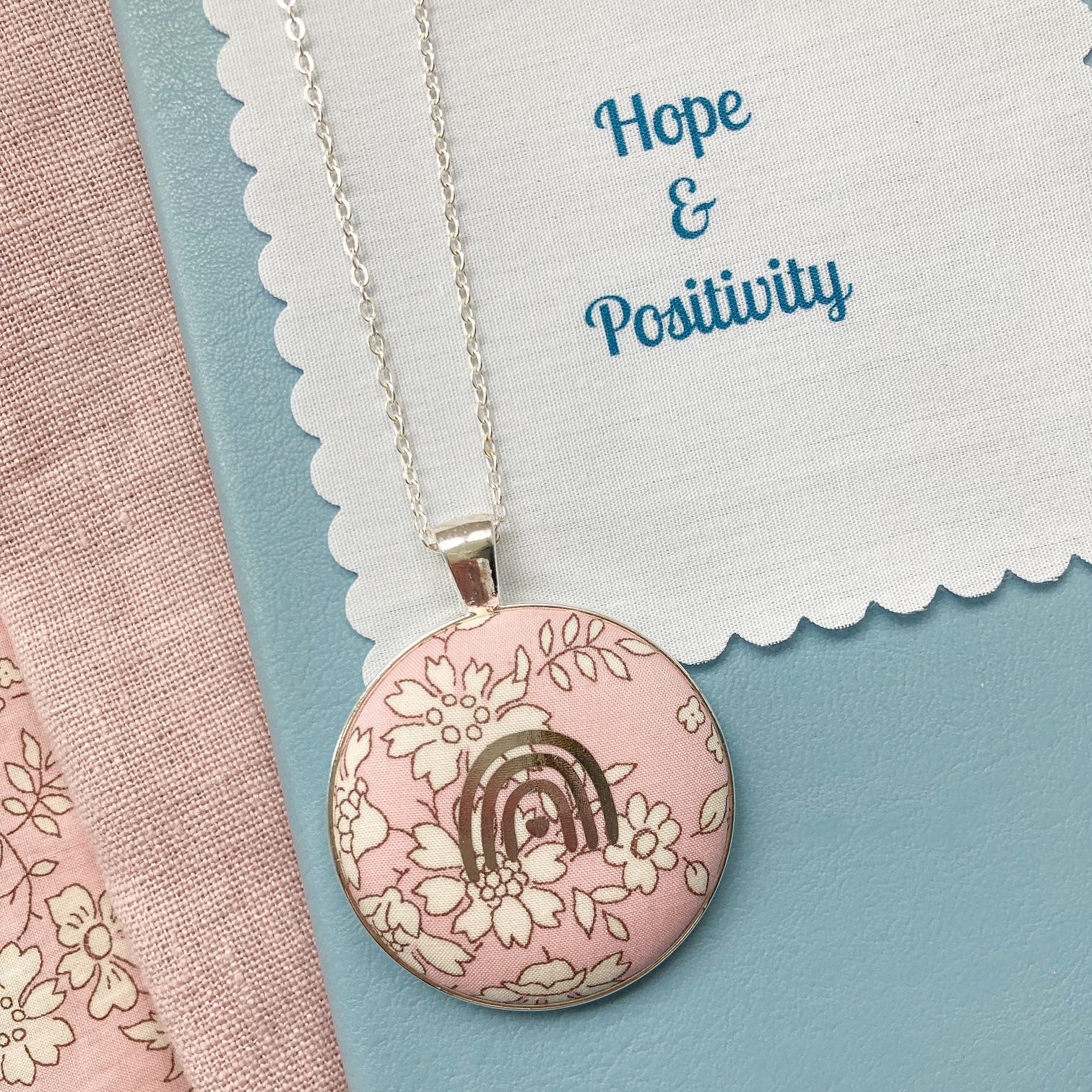 Necklace with a pink pendant featuring a rainbow and flowers on a fabric message with 'Hope & Positivity' text.