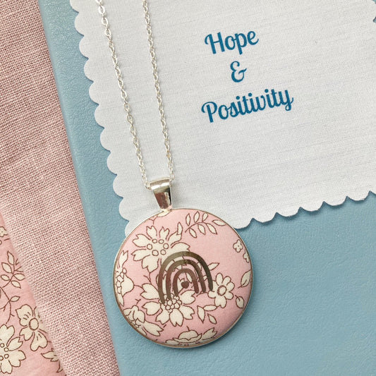 Necklace with a pink pendant featuring a rainbow and flowers on a fabric message with 'Hope & Positivity' text.